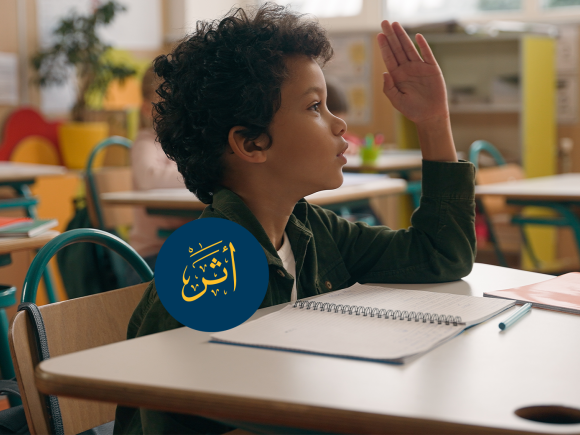 A boy is sitting at a desk in a classroom and raising his hand. Text: أثر