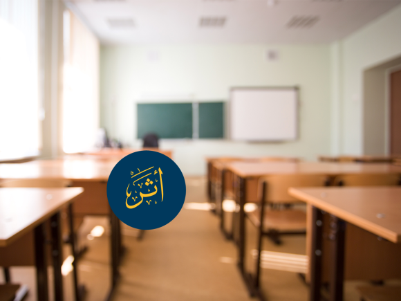 A classroom with empty desks and chairs. There is a blackboard on the wall. Text: أثر