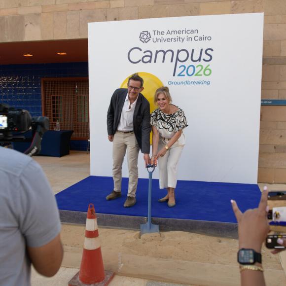 A man and a woman are standing on a step holding a shovel together. Text: Campus 2026 Groundbreaking. The American University in Cairo