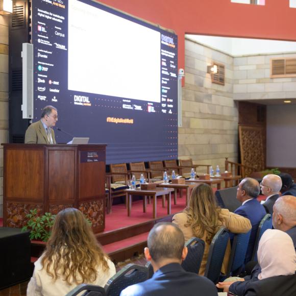 A man is talking into a microphone at a podium. there is a big screen beside him and an audience in front of him