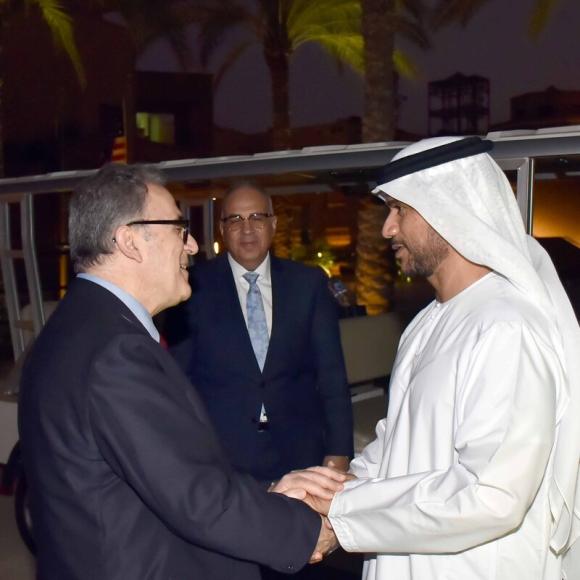 Two men are shaking hands. One is wearing an abaya and headscarf, the other is wearing a suit and glasses. There is a man in the background, he is wearing a suit and glasses. There is a golf cart behind them