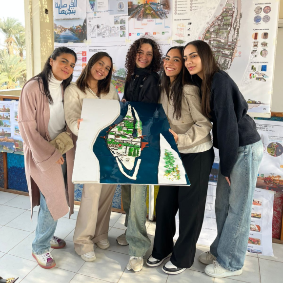 A group of female students smiling and standing together on Qursaya Island, holding an urban design poster the students created