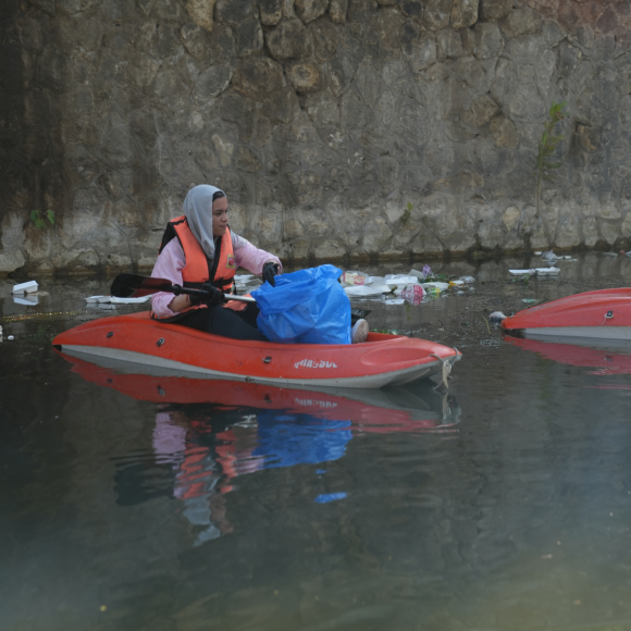 A female student on a boat in the Nile picking up waste from the Nile in a plastic bag