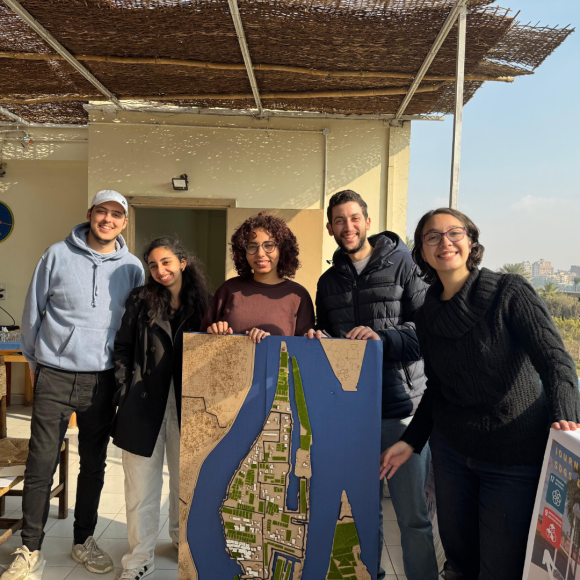 A group of students smiling and standing together on Qursaya Island, holding an urban design poster the students created