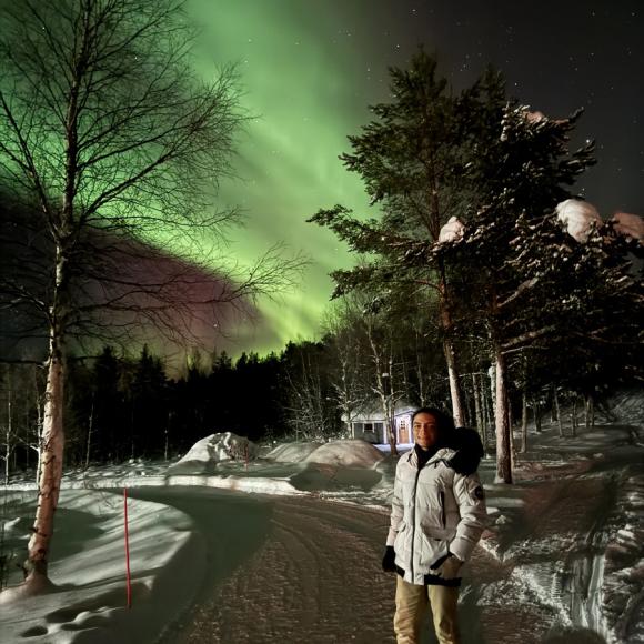 A male is standingin te snowy outdoors with northern lights behind him